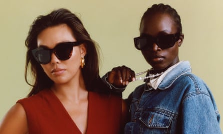 Two models, one wearing a dark red V-neck top and black sunglasses, the other wearing a denim jacket, sunglasses and a pearl necklace which she is pulling out in front of her, both standing in front of a beige background