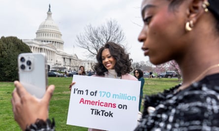 A young Black woman with an iPhone films another Black woman holding a sign that says “I’m 1 of 170 million Americans on TikTok” with the capitol building in the background, on a green lawn.