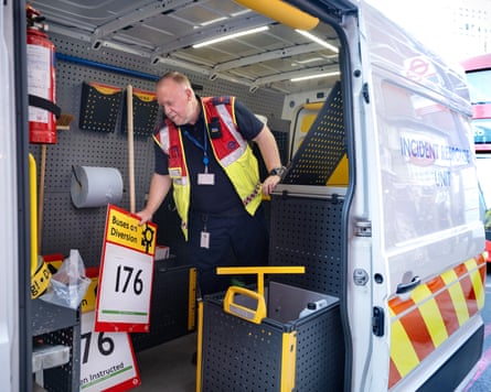 Man in a red and yellow hi-vis vest inside a van, picking up a red and yellow ‘Buses on diversion’ signs.