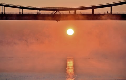 People walking over the bridge are silhouetted against an orange sky, as the rising sun hangs above the river from which mist is rising