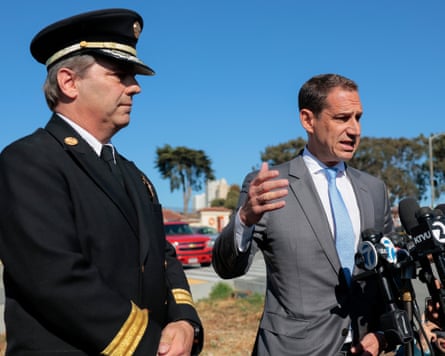 One man in a black vaunted hat stands with hands crossed in front of him as another in a suit speaks and gestures.