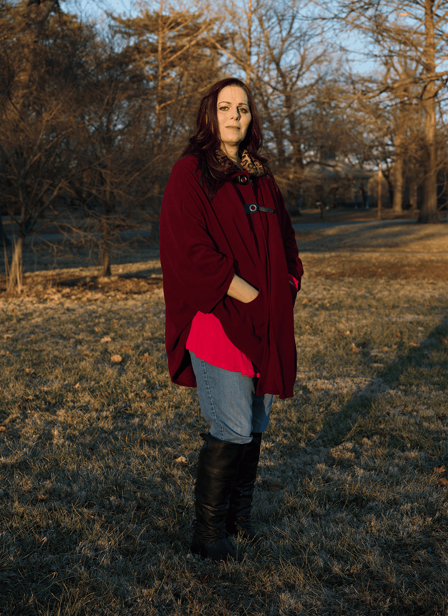 A woman wearing a burgundy cardigan, blue jeans, and black knee high boots stands for a portrait in a park