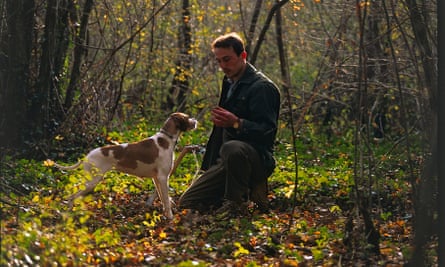 A man kneeling down in the forest holding something, a dog sniffing his hand