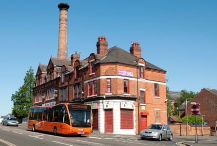 A Trentbarton followed by a car is driven past shops on a Derbyshire road as another car waits to pull out on to the street