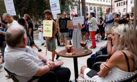 a group of people sit around a cafe table in a street as protesters walk past; one sign reads: your tourism, my misery
