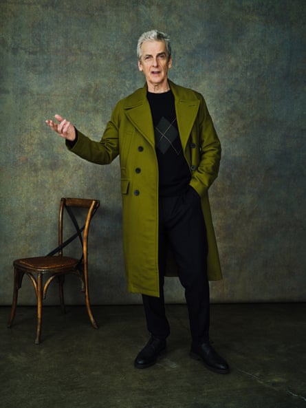 Peter Capaldi, full-length shot, wearing a long olive green coat, undone, hand in black trouser pocket and gesturing with his other hand; also wearing dark jumper with just visible green diamond, and black shoes