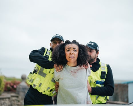 A terrified looking young woman with blood on her face and shirt is apprehended by two police officers on both sides wearing yellow hi-viz jackets