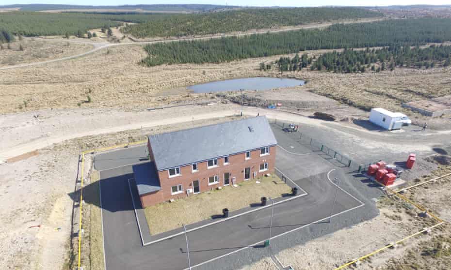A picture of three modern terrace houses with gardens, completely surrounded by a rectangular tarmac road, fenced off, in a sparse hilly landscape