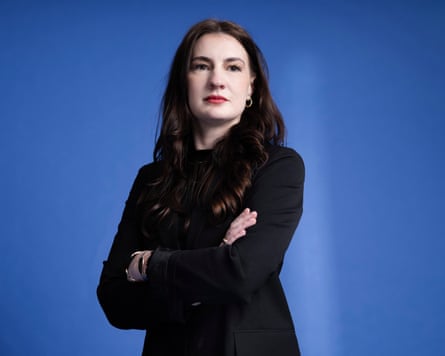 Lucia Osborne-Crowley stands slightly turned away from the camera, her arms folded, with a neutral expression on her face. She has curled dark hair and wears a dark suit