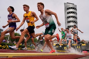 Three runners race beside each other in the foreground as other leap over a water jump behind them. In the background is a skyscraper