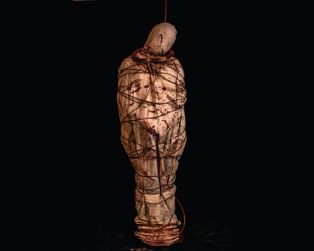 A body wrapped in a bloody shroud and bound with wire that appears to be hanging by a cord from the neck