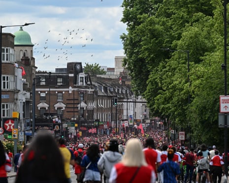 A large crowd fills a street lined with shops and trees, many wearing red and white