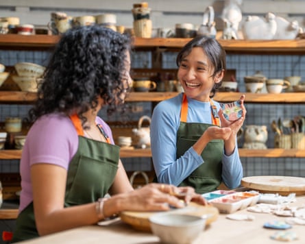 Two women are engaged in a creative pottery session, happily discussing their ceramic piece while seated in a workshop filled with handmade items