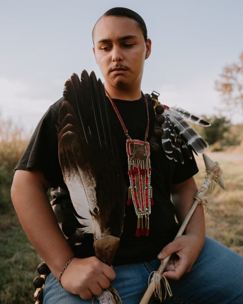 Kaylin Williams holds a feather fan and staff, wearing a beaded pendant necklace outdoors