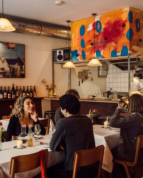 Diners sit at white-clothed tables in a restaurant with an open kitchen, colorful artwork, and exposed ductwork.