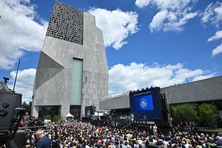 Tall gray building, mostly sunny sky, dozens of people sitting in audience.
