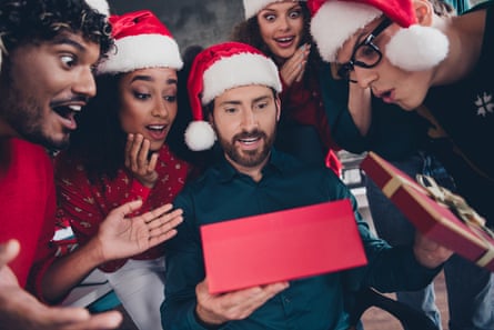 A close up of a group of young people in Santa hats peer in at a young man in Santa hat and dark beard looking at a festive red gift box. Other people are holding boxes