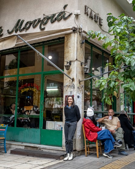 Areti stands outside the cafe. It is a stone corner building with green window and door frames, and its name in green script. She wears a black jumper, black jeans and white Dr Martens boots. There are two people sitting at a table drinking from mugs behind her.