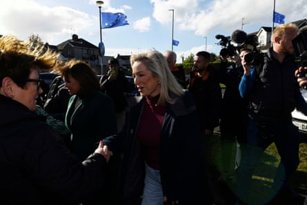 Michelle O'Neill shaking hands with a woman as a media crowd follows behind