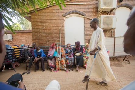 A man with a damaged foot walks with a crutch as other patients wait on a bench at a clinic