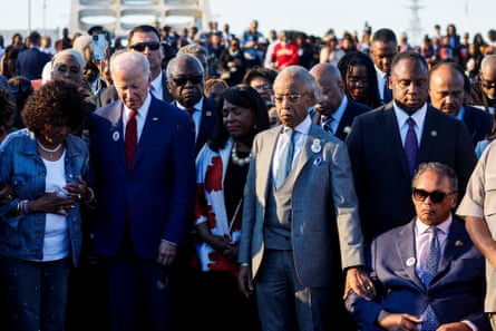 A crowd of mostly Black people, including Rev Al Sharpton, and others, including president Joe Biden, sit and stand on a bridge.
