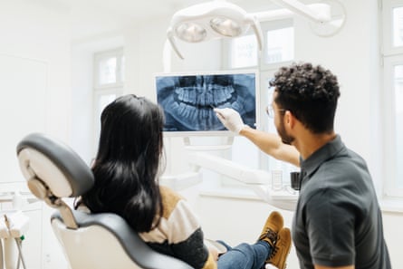An orthodontist gesturing at an x-ray image of one of his patient’s teeth displayed on an overhead monitor as she looks from the dentist chair.