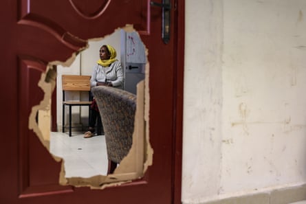A woman in a yellow head scarf sitting in a waiting room photographed through a large hole in a door