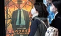 Two women wearing face masks walk by an Oppenheimer film poster