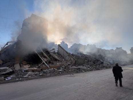 A man walks by a destroyed building that is set against the backdrop of a blue sky, dust still rising from it.