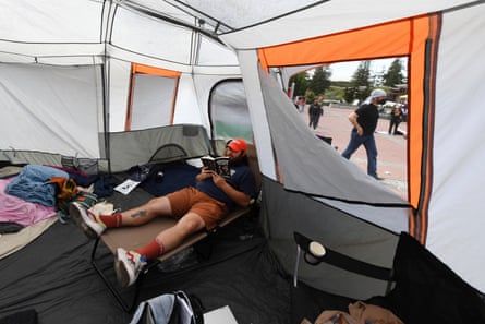 A man lies sprawled on his back inside a tent, with people walking by on a brick plaza visible through the tent flap.