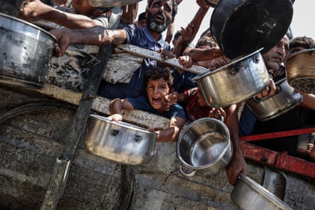 Numerous people including a small child desperately holding out pots and pans from behind a fence