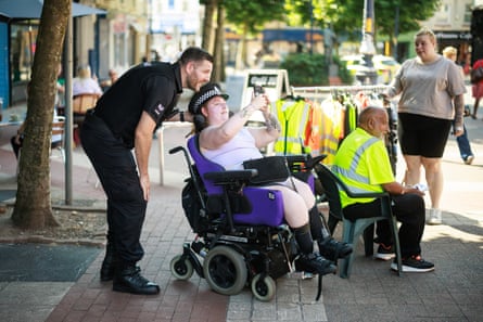 Rob Froggatt leans over a young woman in her 20s who is sitting in a motorised wheelchair and wearing his cap; she is holding up her phone to take a picture of them both, and laughing. He is in a short-sleeved black uniform shirt and has short dark hair and a short-cropped beard. Behind them are a man and a woman beside a rail of hi-vis vests and clothing.