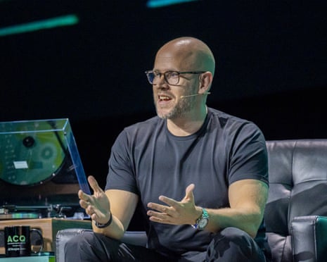 a man in tshirt speaks while seated on stage, a record player beside him