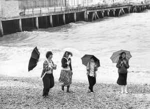 A windy bank holiday at Clacton-on-sea, May 1979. Four women with windswept umbrellas walking along the beach. GNM Archive ref: GUA/6/9/1/1/H