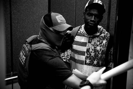 A masked agent holds a man wearing a hoodie bearing a representation of the US flag in an elevator