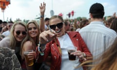 Teenage girl in red leather jacket and sunglasses smiles into cameras as she holds up a lit cigarette and a drink with crowd behind her