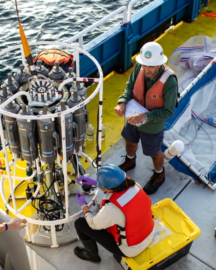 Two people in hard hats standing by a large scientific instrument of 12 scuba-like tanks and pipes in a large frame