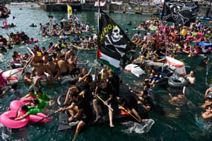 People sit around on makeshift rafts, some of which have ‘skull and crossbones’ pirate flags