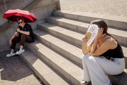 Two women sit on steps in the sun, one under an umbrella, the other using a fan to keep cool