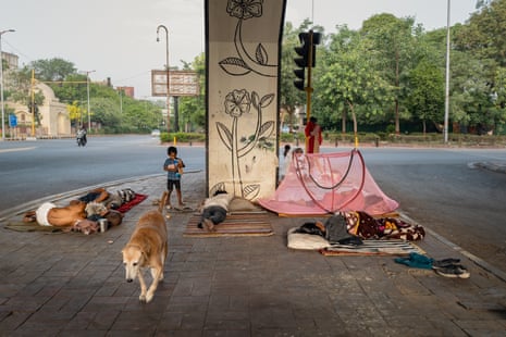 A group of people lie on different mats next to a concrete pillar. A small boy stands looking at a dog which is walking past the sleeping people.