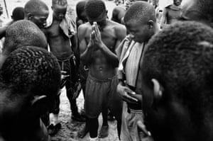 Black and white photo of topless boys standing in a rough circle with their heads bowed in prayer
