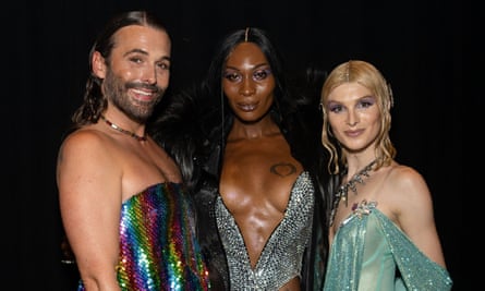 A white man with long hair and a beard, a Black woman with long black hair parted in the middle, and a white woman with long blond hair parted in the middle, wearing sequined, revealing gowns stand in a row, smiling and looking very glamorous.