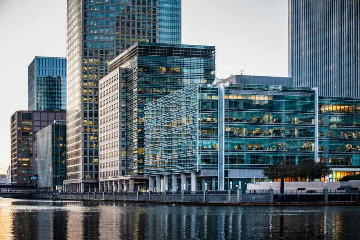 A modern glass-fronted office building in front of taller skyscrapers at a docklands waterside.