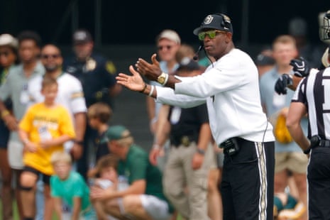 Coach Deion Sanders, in sunglasses and headset, gestures with both hands on the field