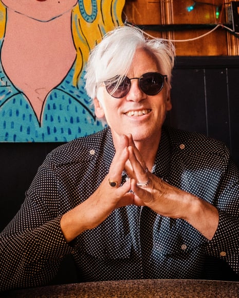 Robyn Hitchcock with white hair and sunglasses sits at a table with hands clasped, in front of a colorful portrait