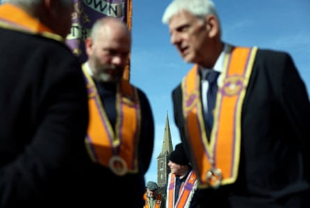 Three members of the Orange Order stand talking, with Drumcree church in the background, under blue skies.