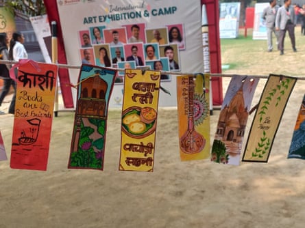 Rectangles of paper decorated with pictures and words hang on a string in front of a sign that reads 'International art exhibition and camp' above photographs of people on colourful backgrounds.