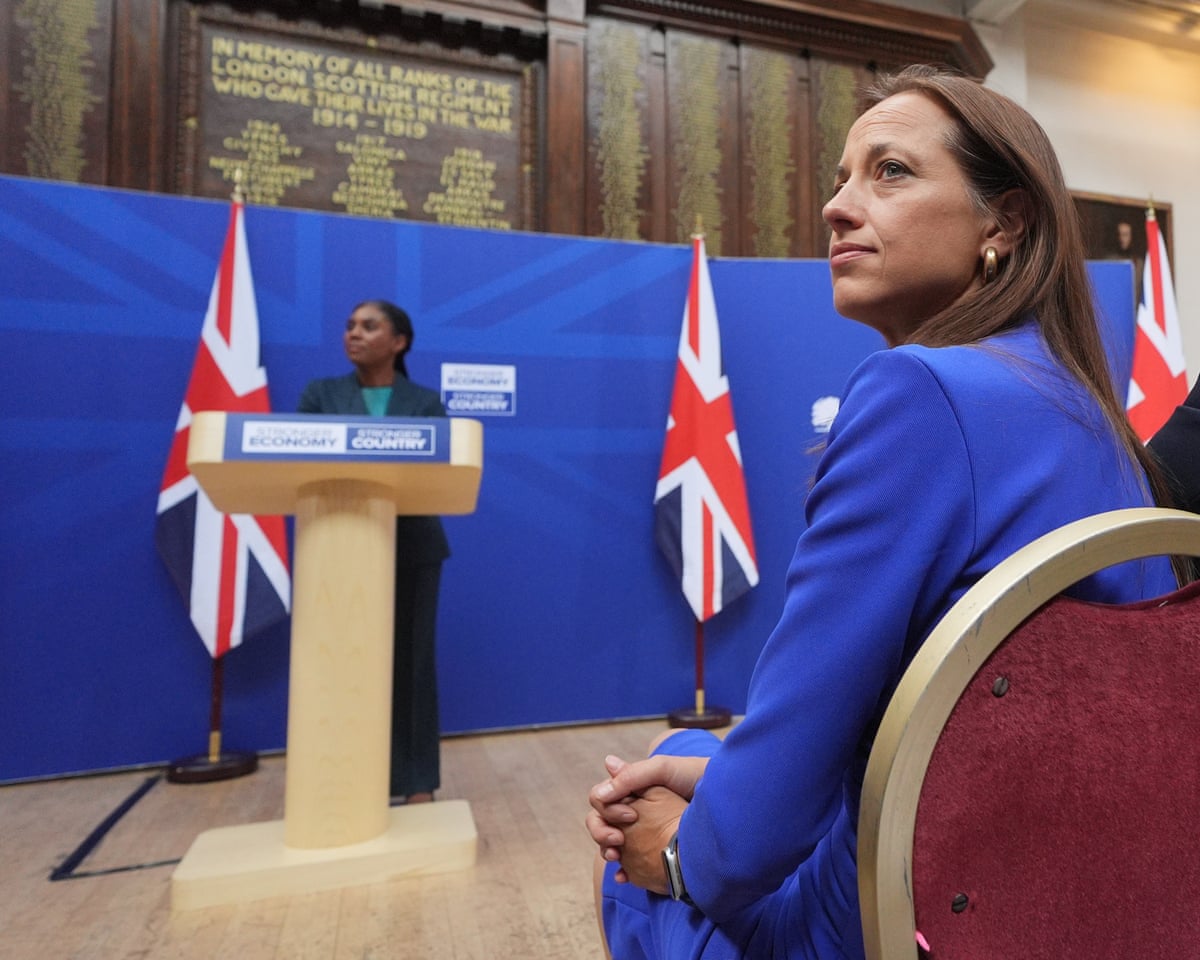 Helen Whately looks to one side as Kemi Badenoch speaks from a lectern.