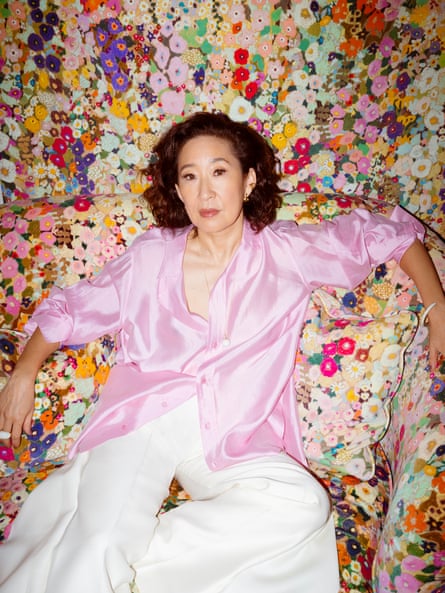 Sandra Oh photographed in pink shirt and white trousers leaning on a floral sofa with a backdrop of floral fabric