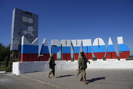 Российские солдаты проходят мимо перекрашенного в цвета российского флага названия города на въезде в Мариуполь в июне.
